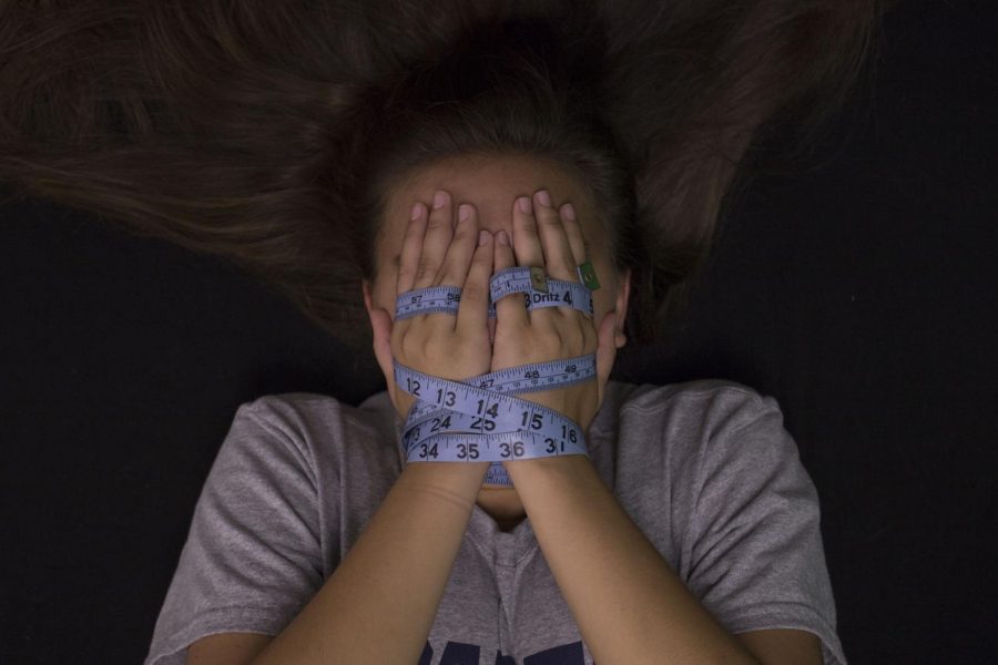 A girls hands are bound in a tape measure as she covers her face. Eating disorders often make individuals feel trapped within their own bodies. 