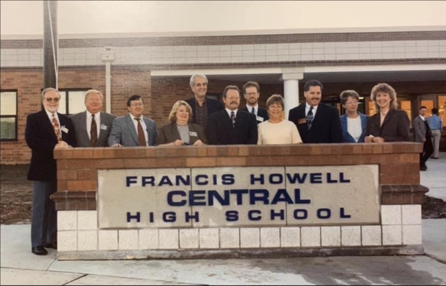 A group of administrators stand smiling at the sign in the front of the building on it’s opening day.