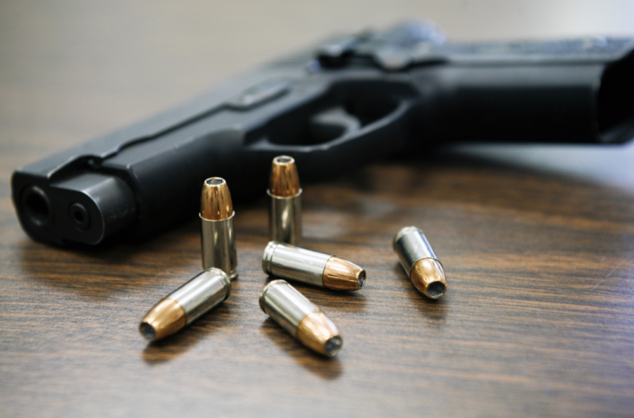 A gun laying on a table surrounded by six bullets. 