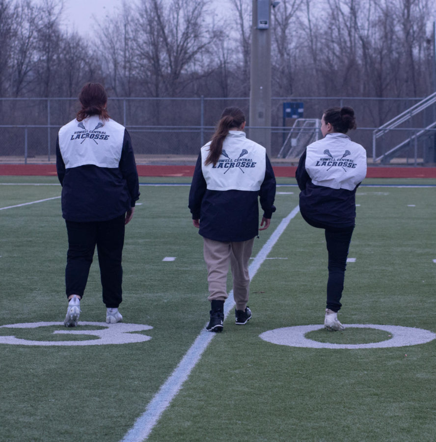 Three of the Varsity Lacrosse players jogging across the stadium field while stretching.