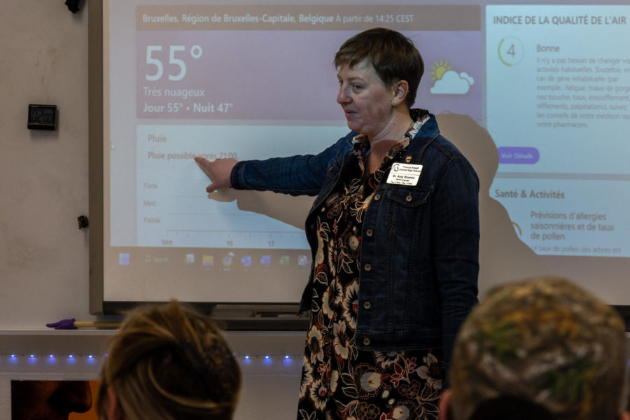 Madame Roznos points to the Smartboard displaying the weather, glancing back at her students.