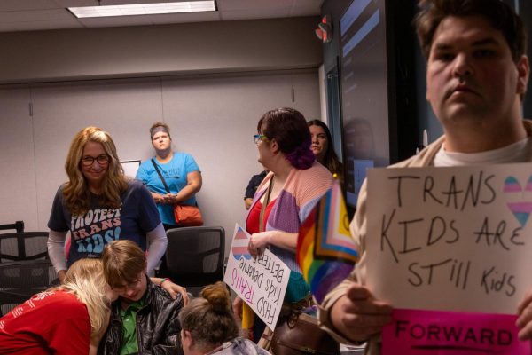 Senior Alexander Collins cries and is consoled after speaking at the podium during the meeting while a previous speaker holds a sign reading TRANS KIDS Are STILLL KIDS.