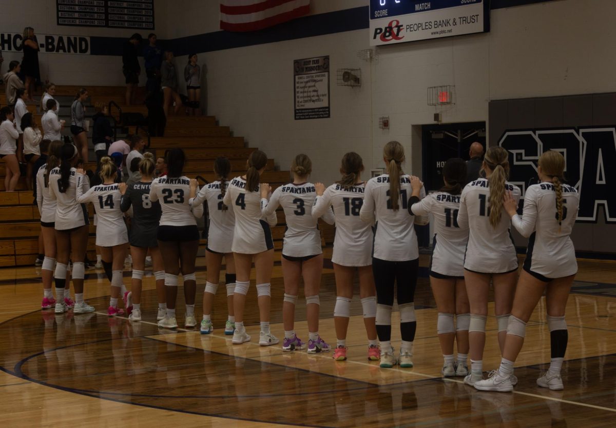 The volleyball team lines up for the national anthem. They lay their hands on each others shoulders, showing themselves as one team.