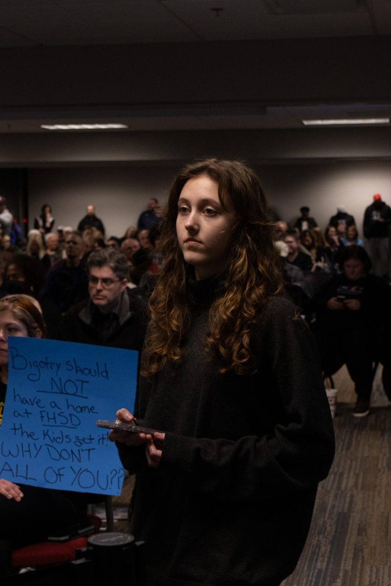 A student stands next to a sign that protests the board decision during the Board of Education meeting. The decision was highly controversial and caused many people to rally against the decision. 