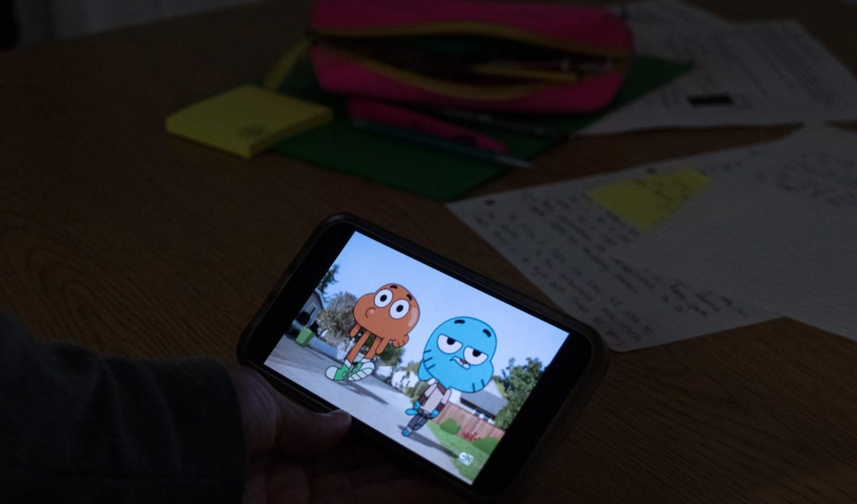 Within a dark room surrounded by papers necessary for studying, a student stares down at their phone, the show The Amazing World of Gumball illuminating them. 