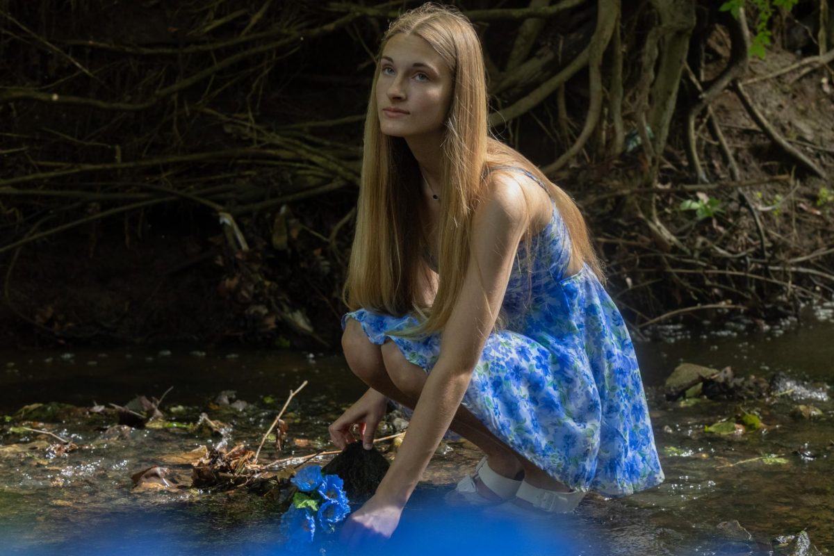 As junior Macey Paniucki picks a blue flower from the creek bed, she looks up, observing the nature around her. The sun shines down on her through the trees, making the water around her glisten in the light.