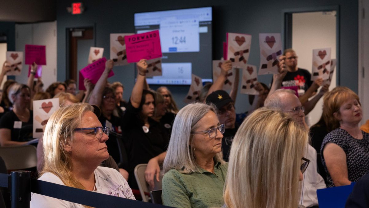 Some meeting attendees sit in silence as many in the background hold signs calling for progress with ideas of unity. The Board meeting proved to be divisive of community members, many making different displays of their beliefs.