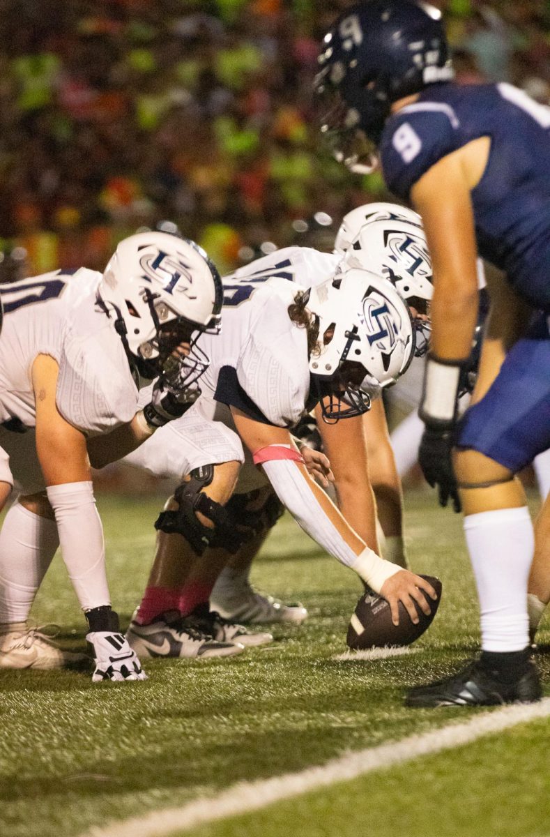 Center junior Nate Gibson, in his junior year, gets ready to snap the ball to his quarterback. He holds the ball firmly on the ground, waiting for the perfect moment to start the play.
