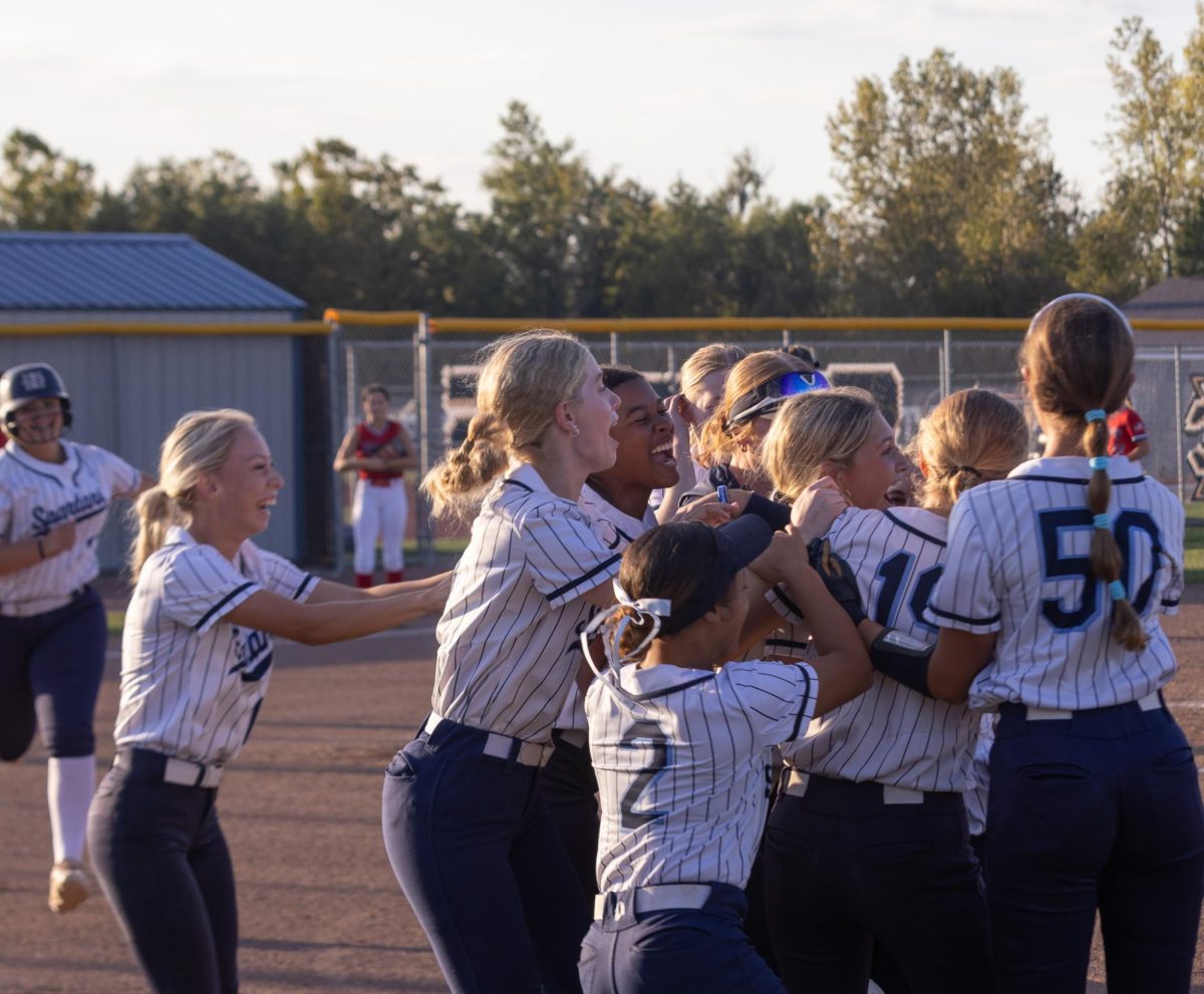 The team rushes to huddle around Callison, jumping and laughing as they congratulate her on her swing and celebrate winning the game together. She won the game for the team and they look forward to playing more games together.