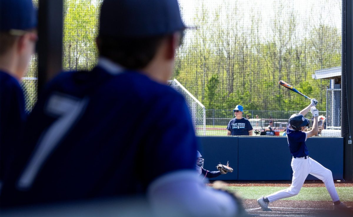 With teammates observing from the dugout, Matt Luczkowski raises the bat high following a strike. After two strikeouts while batting, Luczkowski struck the ball on the third attempt and launched it towards first base. 
