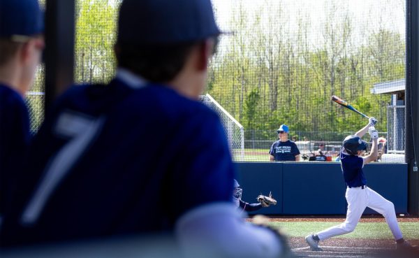 With teammates observing from the dugout, Matt Luczkowski raises the bat high following a strike. After two strikeouts while batting, Luczkowski struck the ball on the third attempt and launched it towards first base. 
