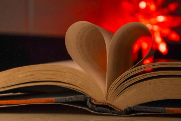 As a book lays open on a table the middle two pages are folded inward to form a heart. The red light in the background shines through the heart to highlight a reader’s love for books. 