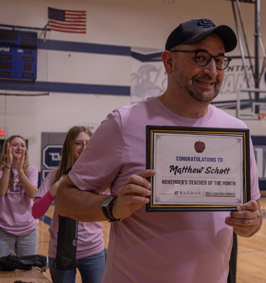 Mr. Matthew Schott smiling while holding his "November Teacher of the Month," plaque awarded to him by St. Louis Post Dispatch. Some of the yearbook staff are congratulating his recognition behind him.