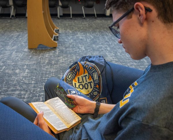 Senior Colin Nichols reading "Fahrenheit 451" by Ray Bradbury in one of the chairs in the Learning commons. He is holding his custom designed Lit Loot bookmark, that of a salamander with the number "451" scrawled across it, and sitting alongside the Lit Loot bag designed by the librarians. 