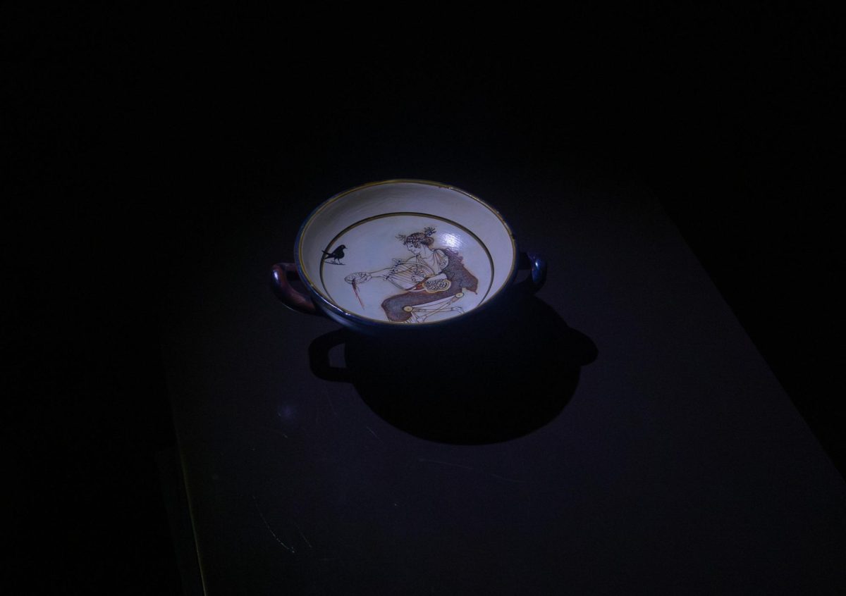 A replica of the Cylix of Apollo—the original of which sits at the Archaeological Museum of Delphi—stands illuminated in a dark room. This drinking cup is a representation of the different outlets used for storytelling.