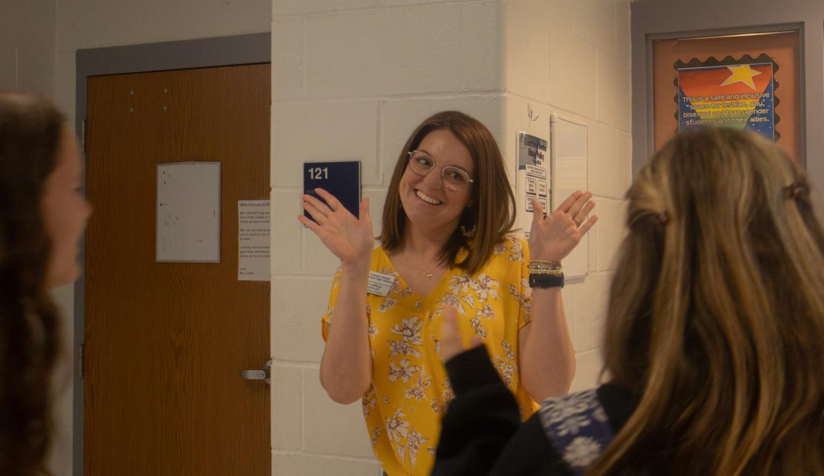 Sarah LaRue greats students into her classroom before the hour starts. The students wave back at her and walk into the class.
